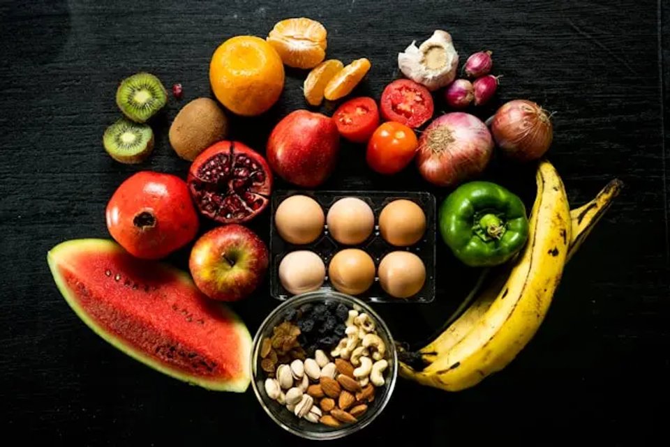 A vibrant arrangement of various fruits and vegetables including watermelon, bananas, apples, kiwis, oranges, tomatoes, and eggs, displayed on a dark surface.