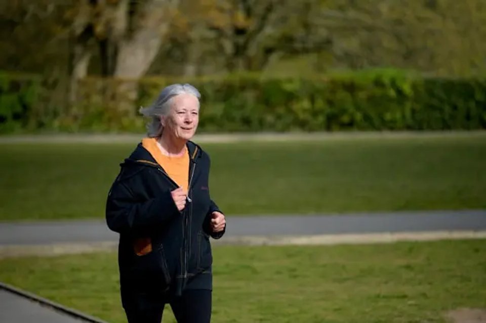 An elderly woman jogging in a park, wearing a black hoodie and smiling as she enjoys her exercise outdoors.