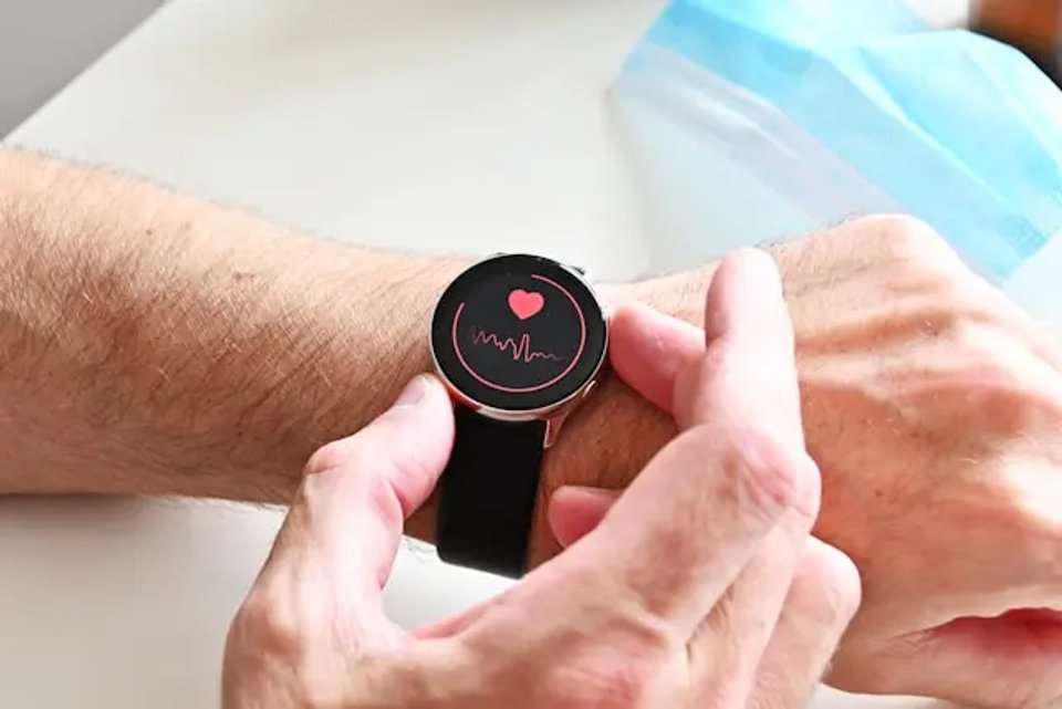 A close-up of a person's wrist wearing a smartwatch displaying a heart rate monitor graphic, with a medical mask in the background.